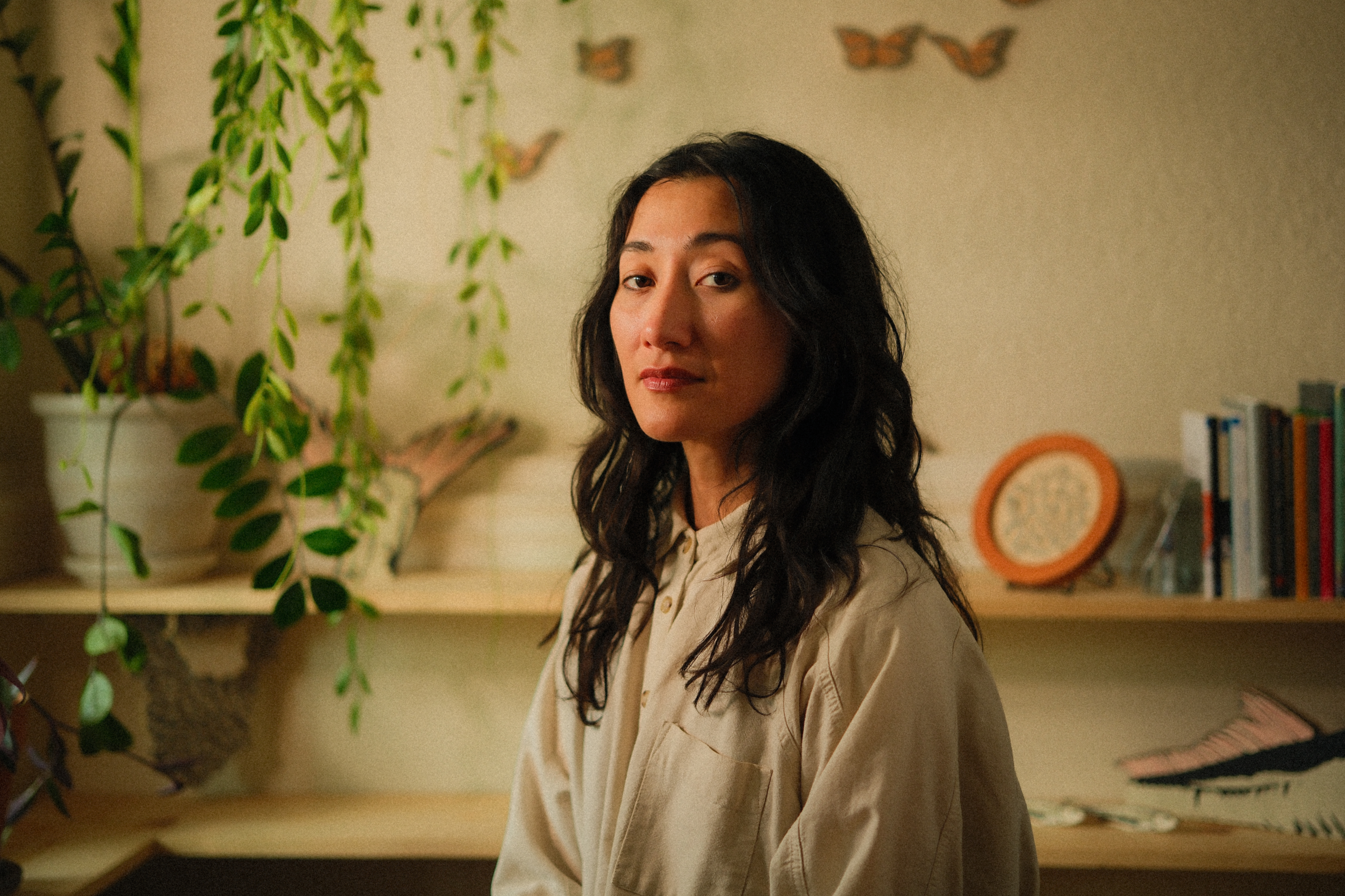 A mixed race woman with long wavy brown hair and olive skin, and brown eyes stares into the camera with a slight smile in her studio. Behind her is a soft blur with shelves of plants, books, and her sculptural woodcut prints