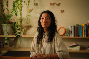 A mixed race woman with long wavy brown hair and olive skin, and brown eyes stares into the camera with a slight smile in her studio. Behind her is a soft blur with shelves of plants, books, and her sculptural woodcut prints
